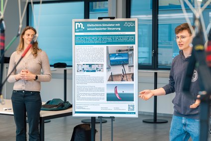 Zwei Personen präsentieren ein Poster „Gleitschirm‑Simulator mit sensorbasierter Steuerung“; Leinen im Vordergrund.
