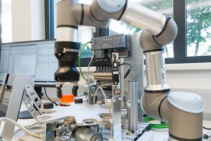 Ein kollaborativer Roboterarm mit Greifer arbeitet über einem Testaufbau auf einem Labortisch; im Hintergrund stehen Steuergeräte, Kabel und ein Monitor vor einem Fenster.
