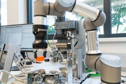 Ein kollaborativer Roboterarm mit Greifer arbeitet über einem Testaufbau auf einem Labortisch; im Hintergrund stehen Steuergeräte, Kabel und ein Monitor vor einem Fenster.