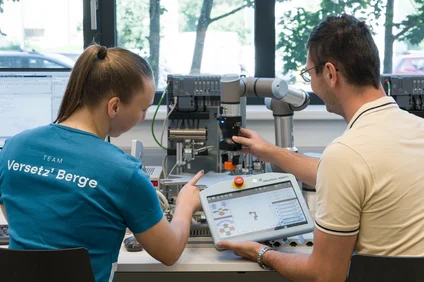 Zwei Personen sitzen an einem Labortisch und bedienen einen kollaborativen Roboterarm; eine Person hält ein Teach-Pendant mit Steueroberfläche, die andere zeigt auf den Versuchsaufbau. Im Hintergrund stehen Steuergeräte, Kabel und ein Fenster; auf einem T‑Shirt steht „Team Versetz’ Berge“.